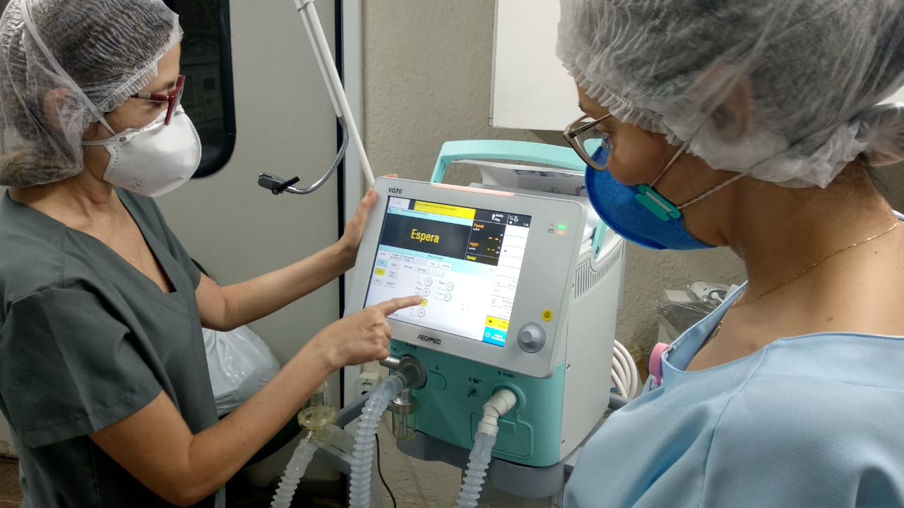 duas mulheres com avental, touca e máscaras cirúrgicos olhando para aparelho respirador entre as duas